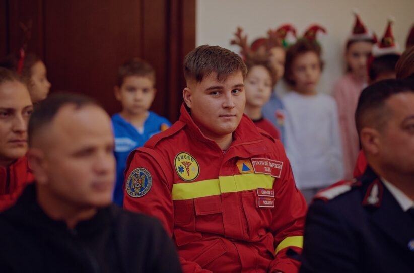 Young man in a red emergency responder uniform sits among a group at an indoor event, patches visible on the chest.
