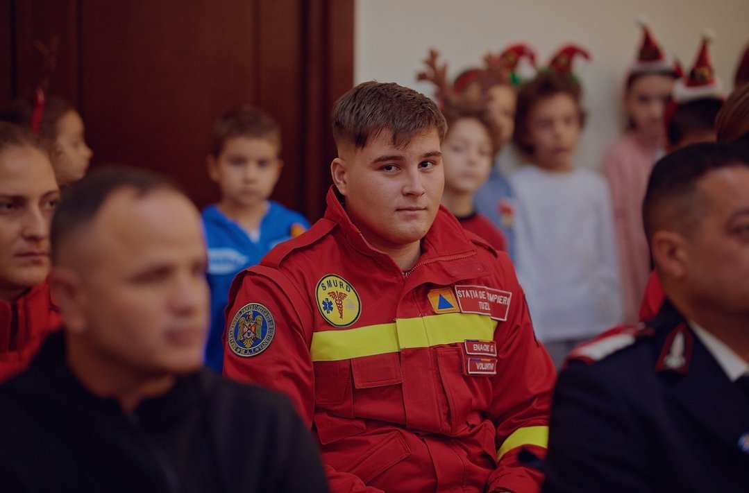 Young man in a red emergency responder uniform sits among a group at an indoor event, patches visible on the chest.