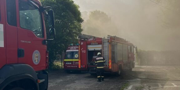 Firefighters in reflective gear connect hoses beside two red fire trucks as thick smoke drifts through a parking lot area