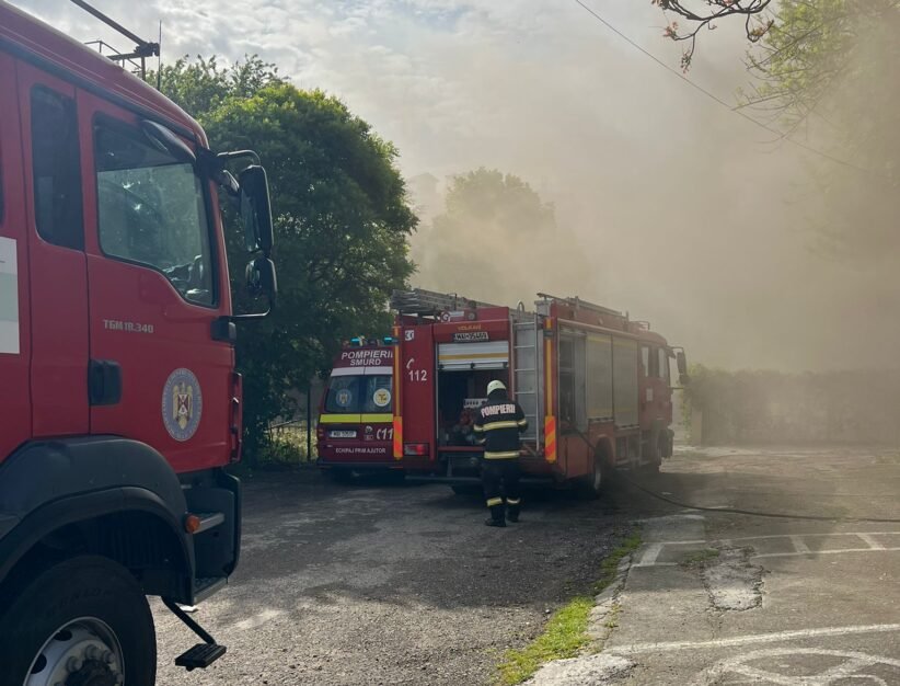 Firefighters in reflective gear connect hoses beside two red fire trucks as thick smoke drifts through a parking lot area