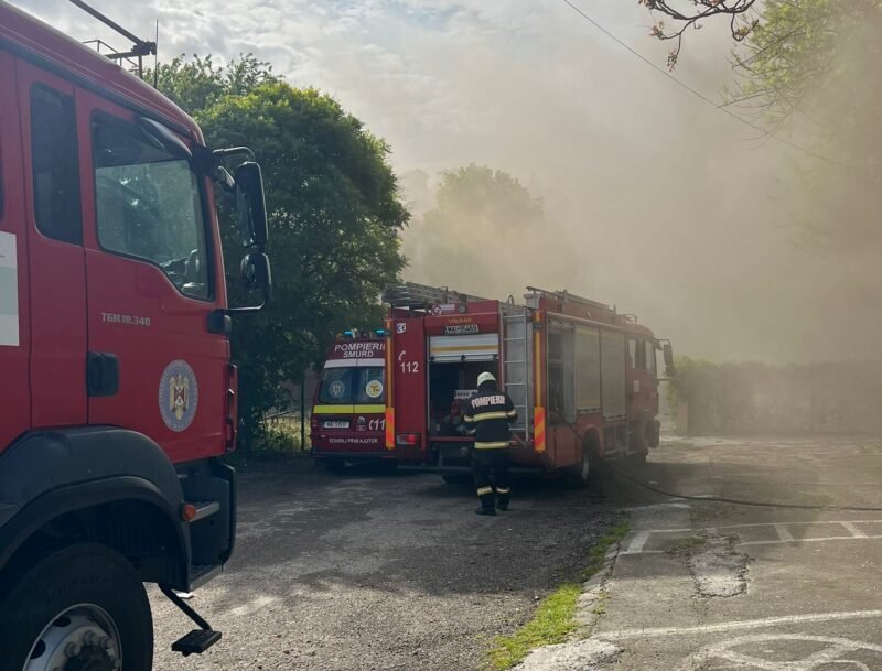Firefighters in reflective gear connect hoses beside two red fire trucks as thick smoke drifts through a parking lot area
