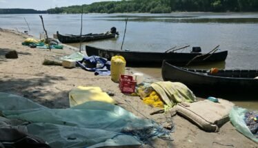 A polluted riverside beach with several wooden boats pulled up on shore and scattered debris, including nets, tarps, and plastic containers. Trees across the water in the background.