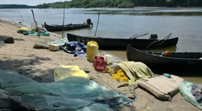 A polluted riverside beach with several wooden boats pulled up on shore and scattered debris, including nets, tarps, and plastic containers. Trees across the water in the background.
