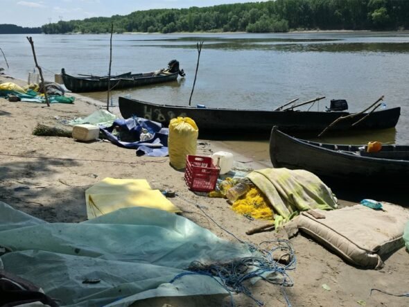 A polluted riverside beach with several wooden boats pulled up on shore and scattered debris, including nets, tarps, and plastic containers. Trees across the water in the background.