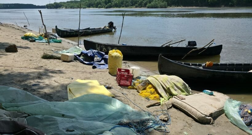 A polluted riverside beach with several wooden boats pulled up on shore and scattered debris, including nets, tarps, and plastic containers. Trees across the water in the background.