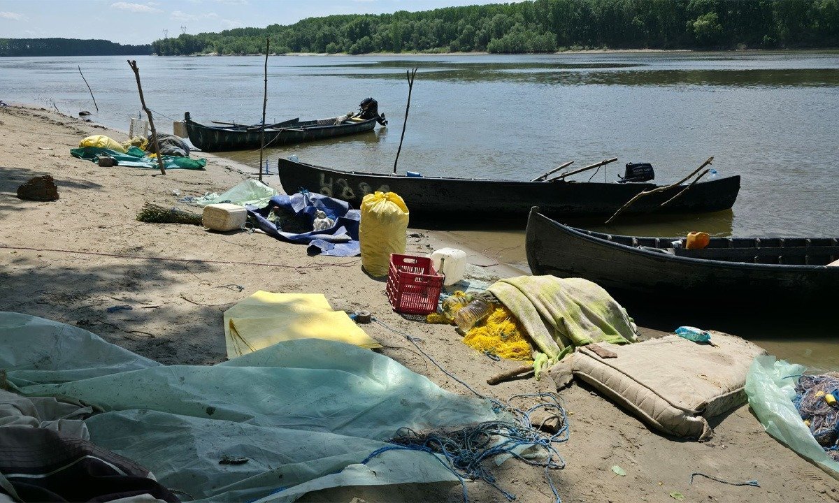 A polluted riverside beach with several wooden boats pulled up on shore and scattered debris, including nets, tarps, and plastic containers. Trees across the water in the background.