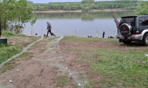 Person walking toward a riverbank with fishing gear laid out on the ground; a vehicle parked to the right by the shore.