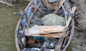 A weathered wooden boat cluttered with plastic bottles, fishing nets, and bags, stranded in murky water near a muddy shoreline.