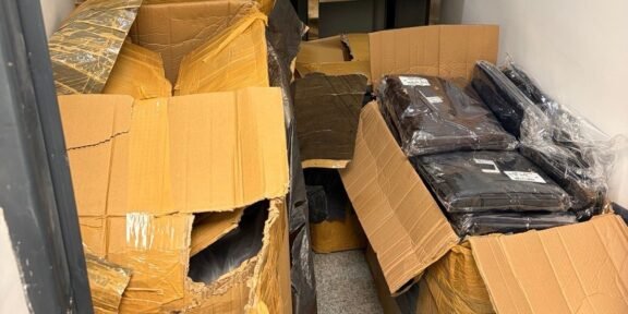 Overflowing storage closet with torn cardboard boxes and sealed black bags.