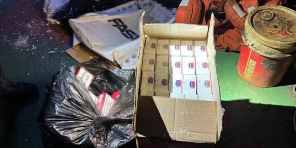 Open cardboard box filled with many small white boxes with blue and red labels, surrounded by bags of trash and a red metal drum on a dirty floor.