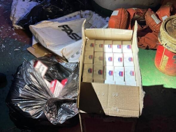 Open cardboard box filled with many small white boxes with blue and red labels, surrounded by bags of trash and a red metal drum on a dirty floor.