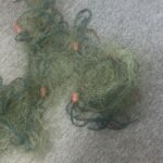 Tangled green fishing net with blue lines and orange floats on a gray carpet. A plastic bag sits nearby for scale.