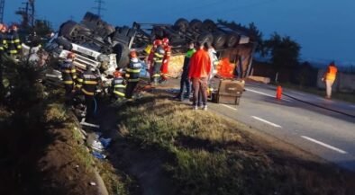 Overturned truck on a highway with emergency responders and a few bystanders at dusk/early evening scene of a crash site visualized by flashing gear and safety equipment, road cones nearby.