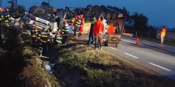 Overturned truck on a highway with emergency responders and a few bystanders at dusk/early evening scene of a crash site visualized by flashing gear and safety equipment, road cones nearby.