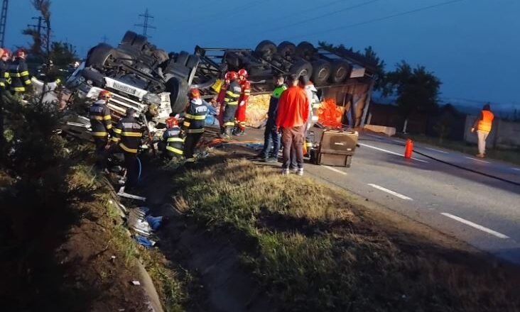 Overturned truck on a highway with emergency responders and a few bystanders at dusk/early evening scene of a crash site visualized by flashing gear and safety equipment, road cones nearby.