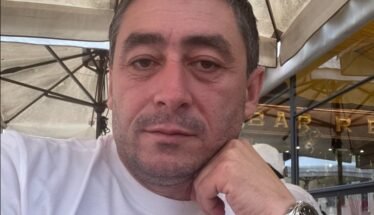 Close-up of a man in a white shirt resting his chin on his hand at an outdoor cafe, wearing a black strap watch.
