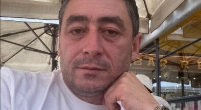 Close-up of a man in a white shirt resting his chin on his hand at an outdoor cafe, wearing a black strap watch.
