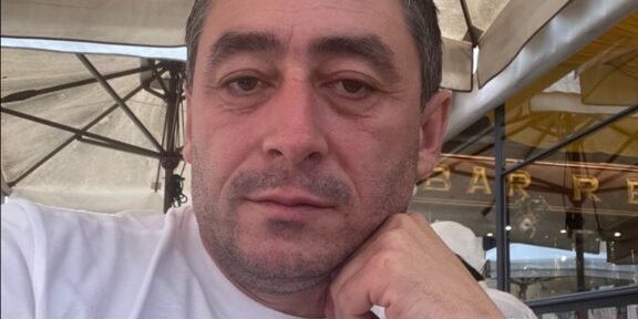 Close-up of a man in a white shirt resting his chin on his hand at an outdoor cafe, wearing a black strap watch.