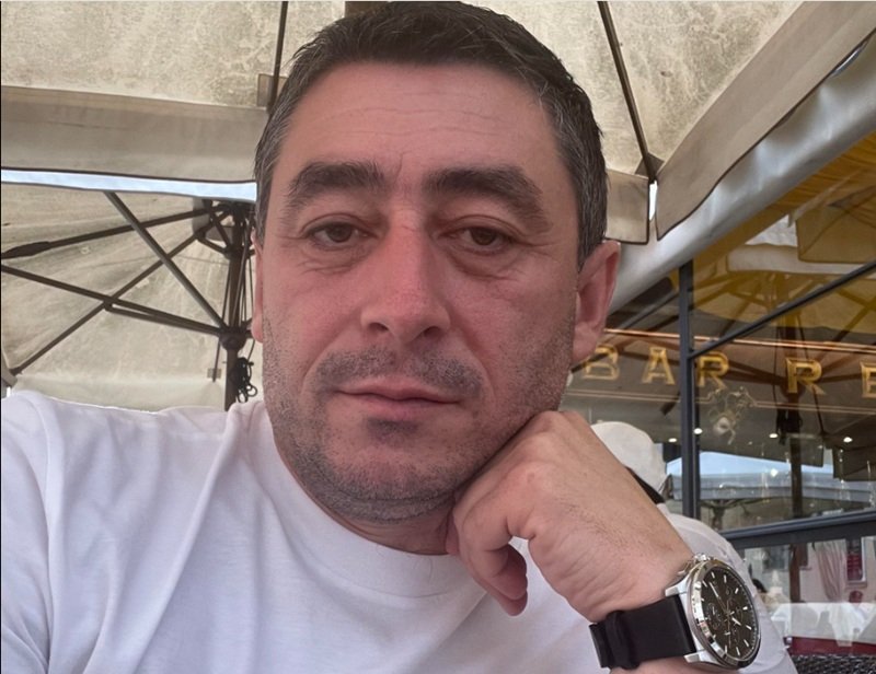 Close-up of a man in a white shirt resting his chin on his hand at an outdoor cafe, wearing a black strap watch.
