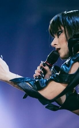Female singer in a black leather outfit extends her hands toward the audience while singing into a microphone on stage.