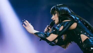 Female singer in a black leather outfit extends her hands toward the audience while singing into a microphone on stage.