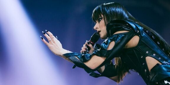 Female singer in a black leather outfit extends her hands toward the audience while singing into a microphone on stage.