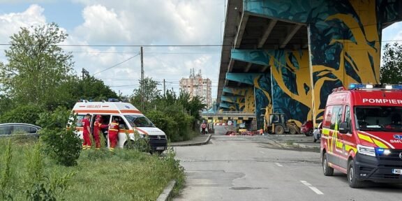 Emergency responders in red uniforms stand beside an ambulance and a red fire service vehicle under a graffiti-covered elevated highway.