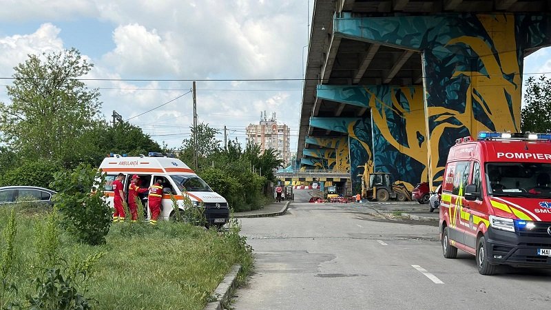 Emergency responders in red uniforms stand beside an ambulance and a red fire service vehicle under a graffiti-covered elevated highway.