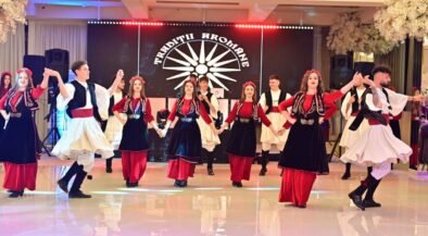 Dancers in red, black, and white traditional outfits perform a coordinated folk dance on a stage with a sunburst backdrop.