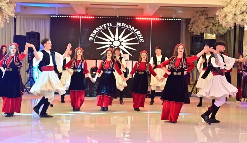 Dancers in red, black, and white traditional outfits perform a coordinated folk dance on a stage with a sunburst backdrop.