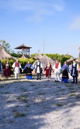Group of people in traditional clothing form a circle outdoors, holding hands, with thatched huts in a sunny setting (cultural gathering).