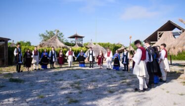 Group of people in traditional clothing form a circle outdoors, holding hands, with thatched huts in a sunny setting (cultural gathering).