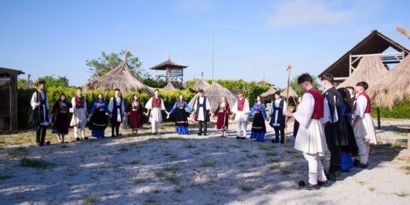 Group of people in traditional clothing form a circle outdoors, holding hands, with thatched huts in a sunny setting (cultural gathering).