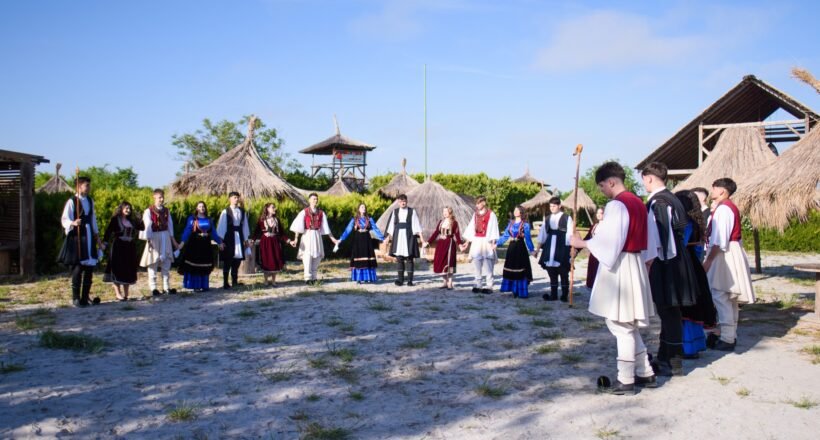 Group of people in traditional clothing form a circle outdoors, holding hands, with thatched huts in a sunny setting (cultural gathering).
