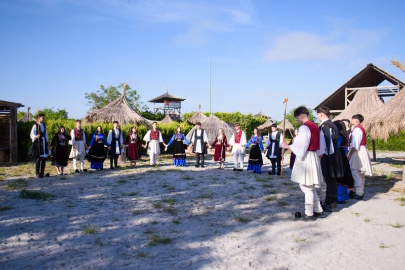 Group of people in traditional clothing form a circle outdoors, holding hands, with thatched huts in a sunny setting (cultural gathering).
