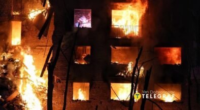 A brick building is engulfed in bright orange flames at night, with multiple windows burning and smoke filling the scene.