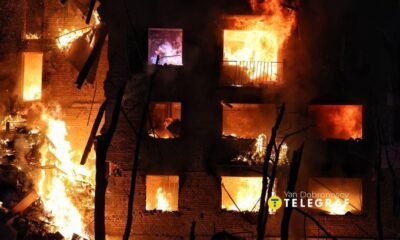 A brick building is engulfed in bright orange flames at night, with multiple windows burning and smoke filling the scene.