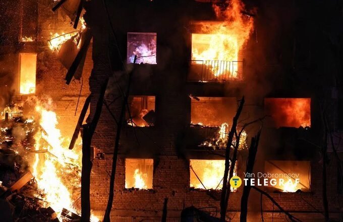 A brick building is engulfed in bright orange flames at night, with multiple windows burning and smoke filling the scene.