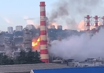 Industrial factory with a tall red-and-white smokestack on fire, flames and thick smoke rising above the plant.