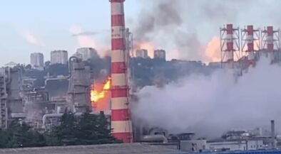 Industrial factory with a tall red-and-white smokestack on fire, flames and thick smoke rising above the plant.