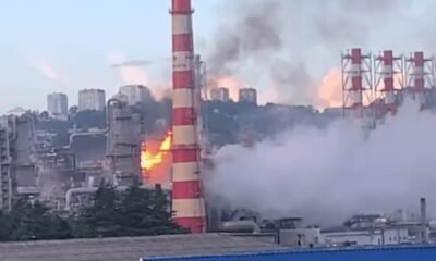 Industrial factory with a tall red-and-white smokestack on fire, flames and thick smoke rising above the plant.