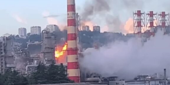Industrial factory with a tall red-and-white smokestack on fire, flames and thick smoke rising above the plant.