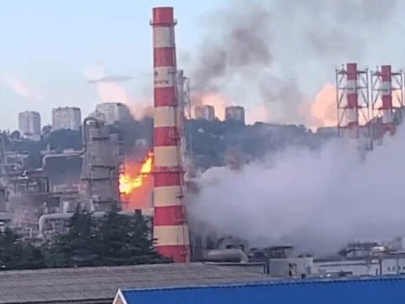 Industrial factory with a tall red-and-white smokestack on fire, flames and thick smoke rising above the plant.