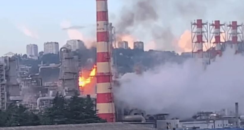 Industrial factory with a tall red-and-white smokestack on fire, flames and thick smoke rising above the plant.