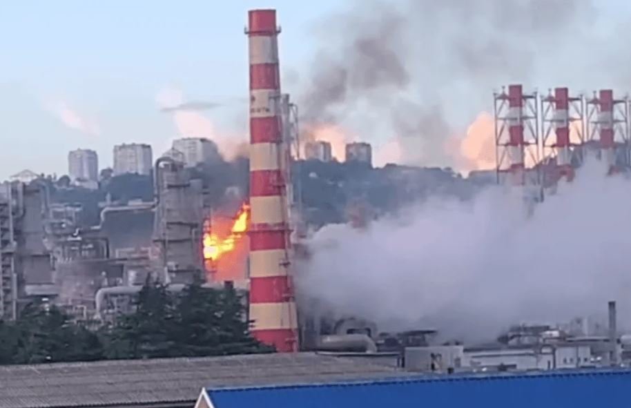 Industrial factory with a tall red-and-white smokestack on fire, flames and thick smoke rising above the plant.