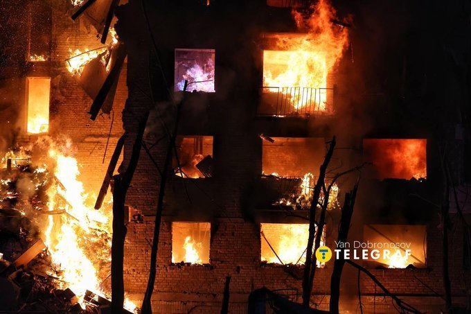 A brick building is engulfed in bright orange flames at night, with multiple windows burning and smoke filling the scene.