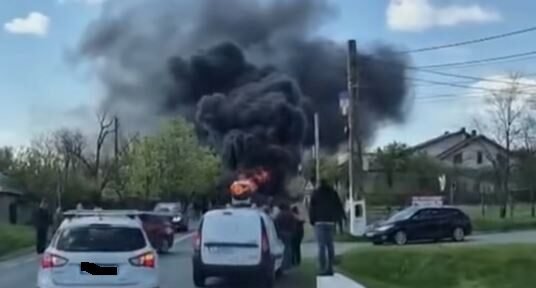 Car on a two-lane road is on fire, sending a large plume of black smoke while bystanders watch and other cars stop nearby on a residential street.