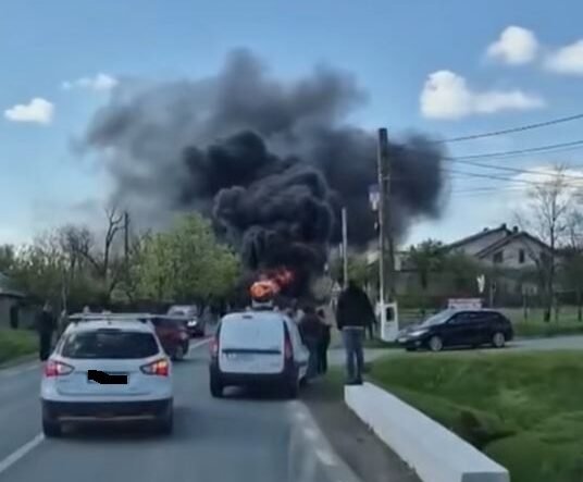 Car on a two-lane road is on fire, sending a large plume of black smoke while bystanders watch and other cars stop nearby on a residential street.