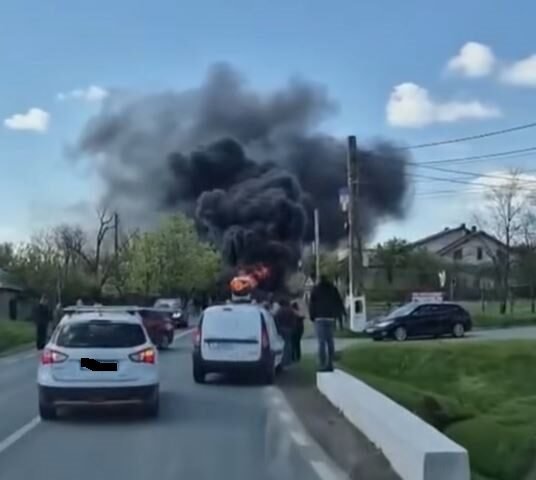 Car on a two-lane road is on fire, sending a large plume of black smoke while bystanders watch and other cars stop nearby on a residential street.