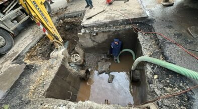 Construction site: a deep excavation with water in the pit, a worker standing inside wearing a blue hard hat/vest, and large hoses looping in and out of the trench.